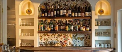 A bar area with shelves of liquor bottles, glasses, and a decorative floral wall.