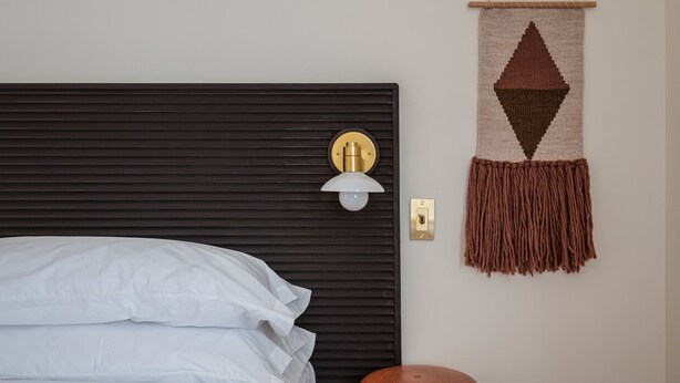 A bed with white linens, a wall-mounted light fixture, a doorbell, and a decorative wall hanging. - Eastwind Hotel Lake Placid (Lake Placid)