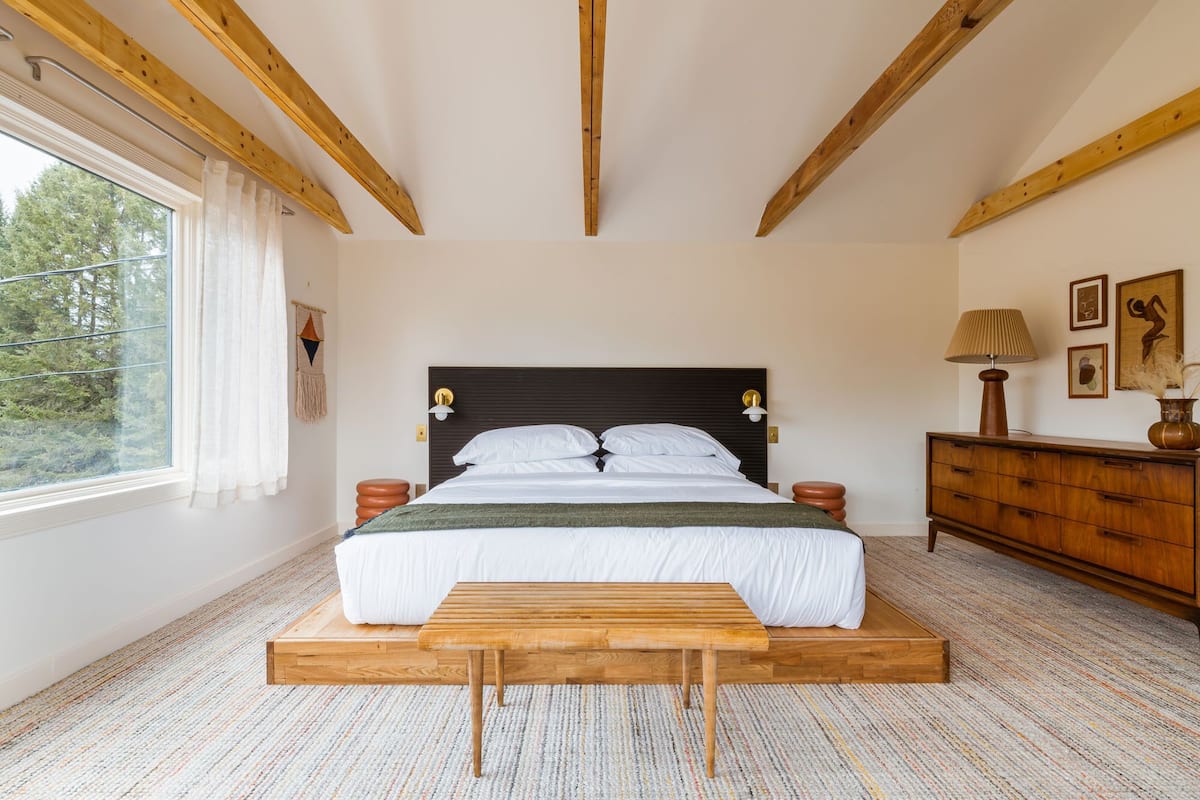 A bedroom with a wooden bed frame, a bench at the foot of the bed, a wooden dresser, and a window with curtains.