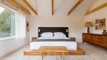 Un dormitorio con estructura de cama de madera, un banco al pie de la cama, cómoda de madera y una ventana con cortinas.