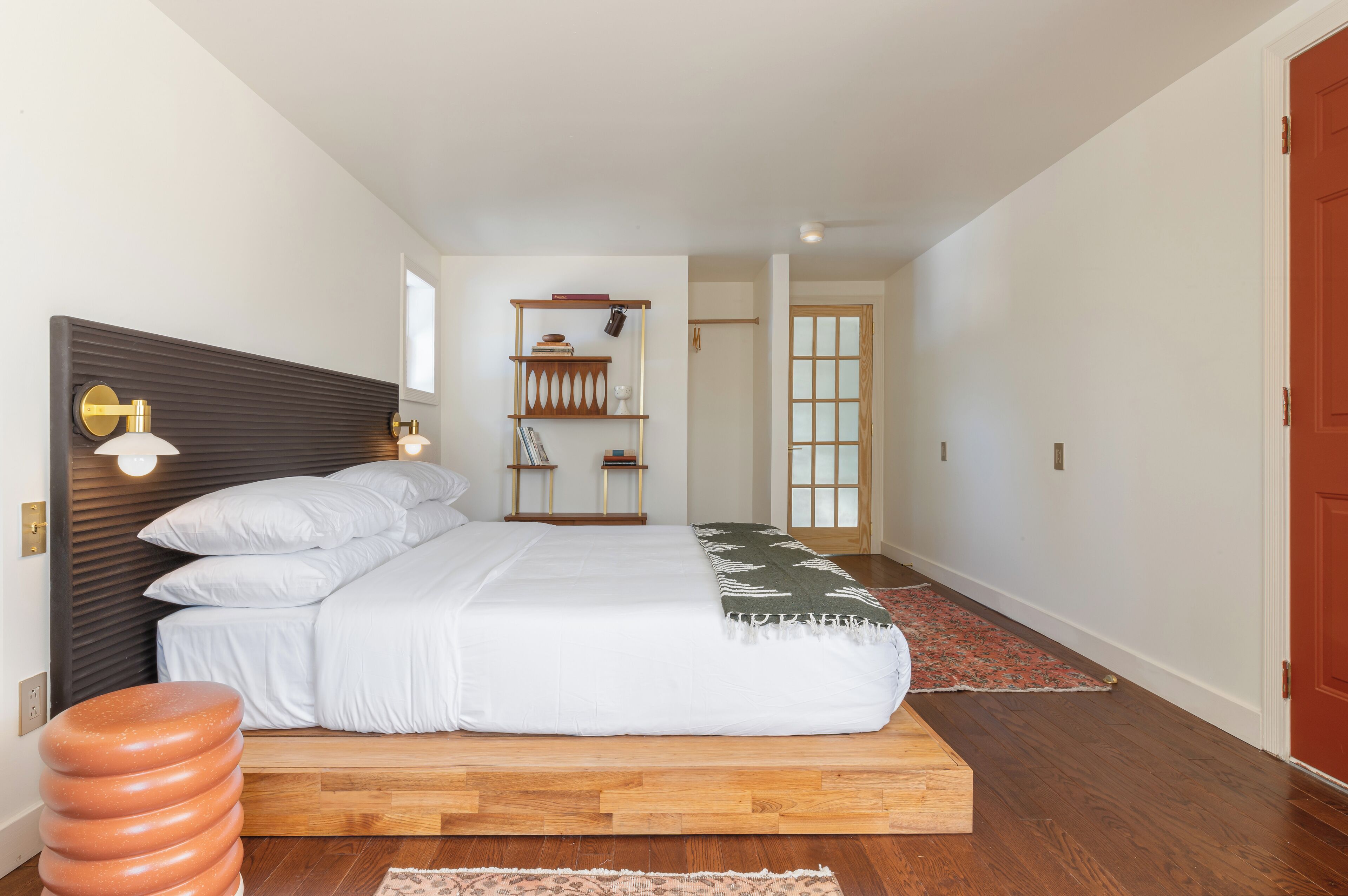 Un dormitorio con una cama plataforma de madera, cabecera con iluminación integrada, mesita de noche y velador. Se ve una ventana con vistas al exterior, una puerta y un estante pequeño con libros y objetos decorativos.