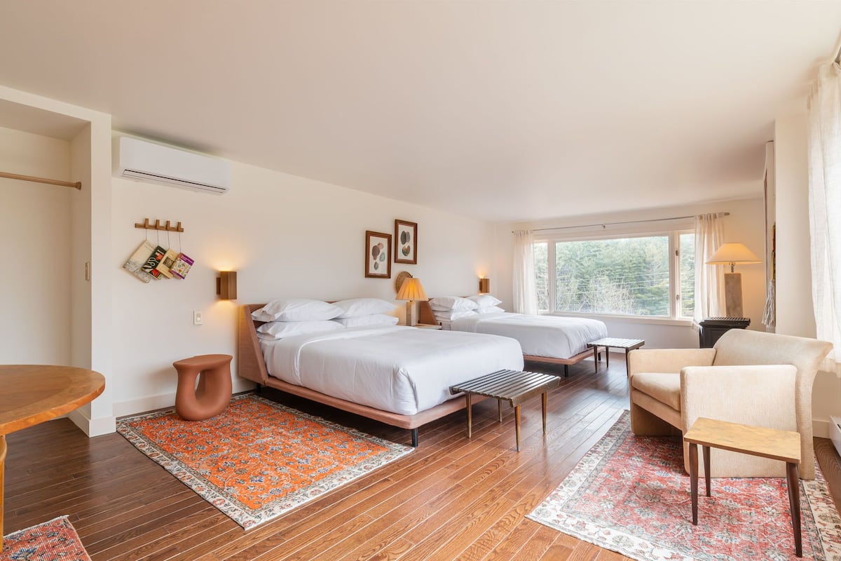 A hotel room with two beds, a wooden table, a chair, and a window with a view of trees.