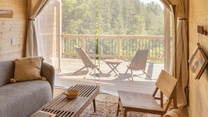 Ein gemütliches Blockhaus-Interieur mit Holzbalkendecke, einer Couch, einem Tisch und zwei Stühlen auf einem Balkon mit Blick auf Bäume.