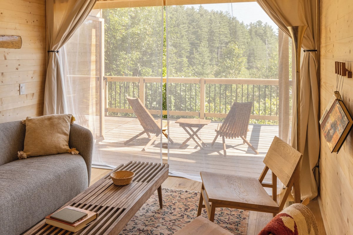 A cozy cabin interior with a wooden ceiling, a sofa, a table, and two chairs on a balcony with a view of trees.
