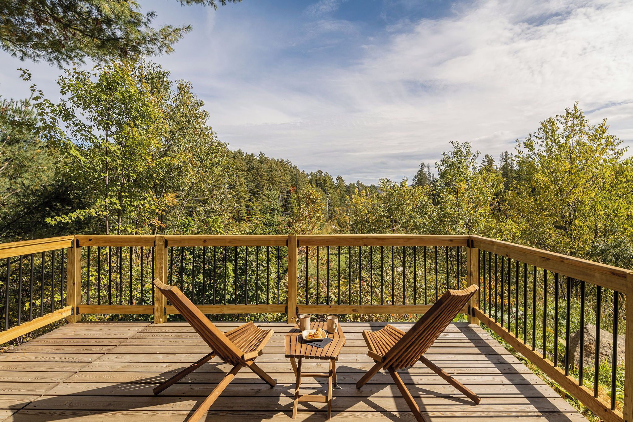 A wooden deck with two wooden chairs and a small table, surrounded by a railing, overlooking a forested area.