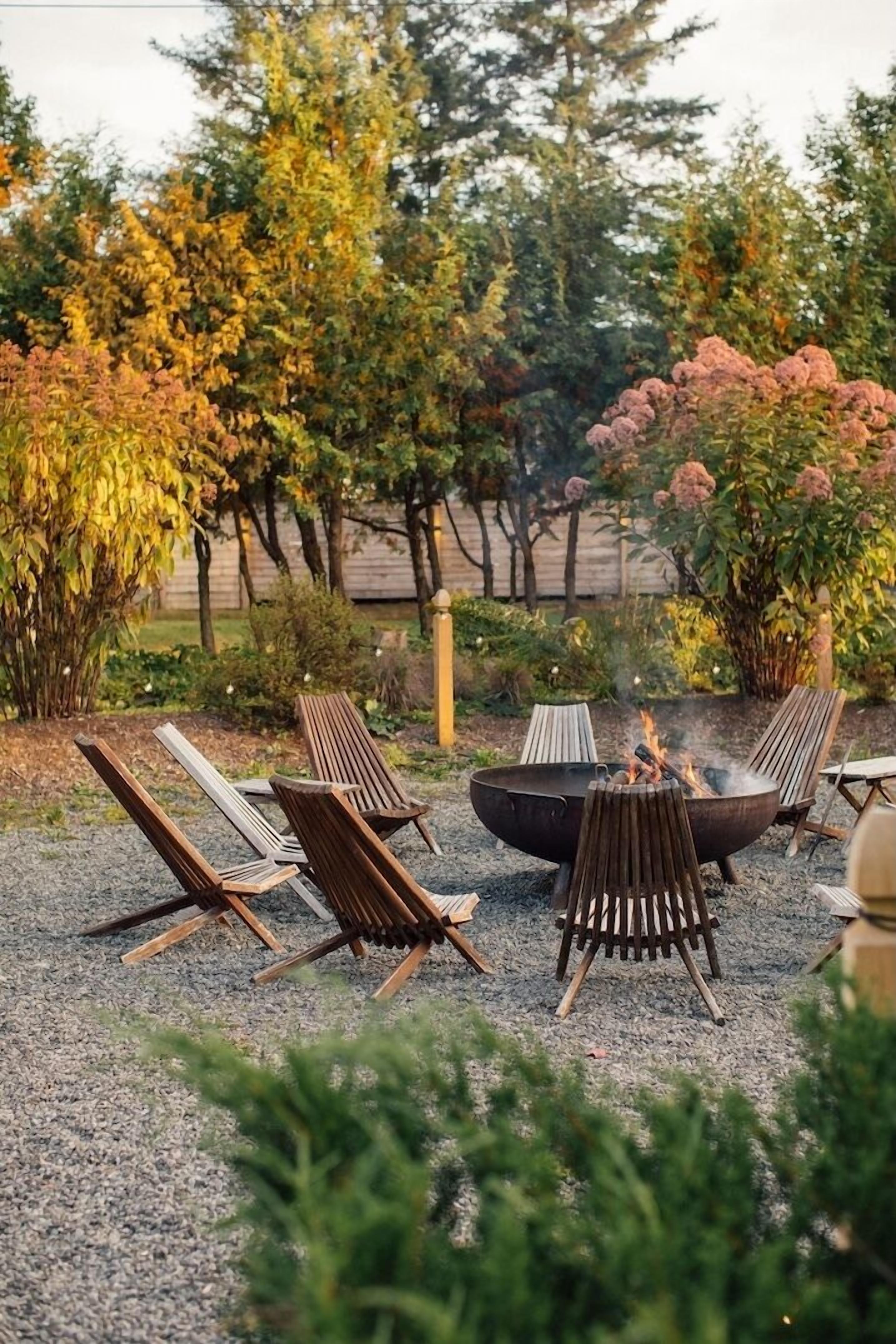 Een vuurkorf omringd door vier lounge stoelen in een tuin met bomen en struiken.