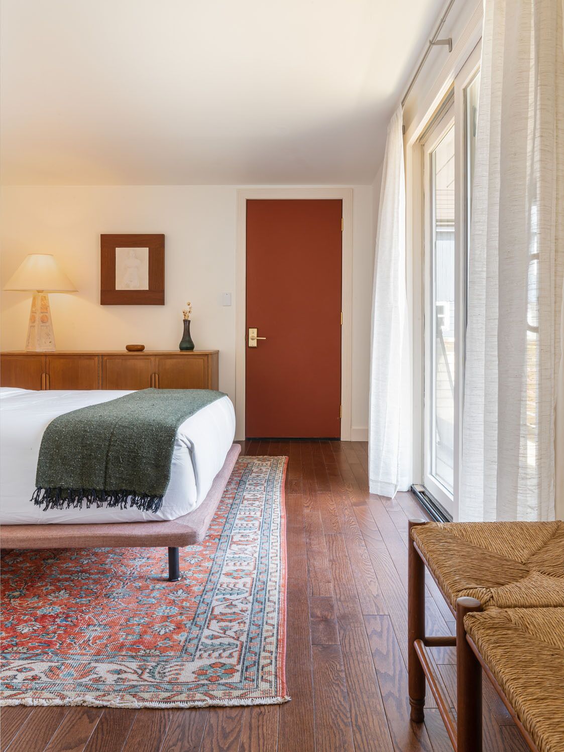 Habitación de hotel con una cama, una mesita de noche de madera, una lámpara, un cuadro enmarcado, una puerta y una ventana con cortinas.