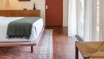 Habitación de hotel con una cama, una mesita de noche de madera, una lámpara, un cuadro enmarcado, una puerta y una ventana con cortinas.