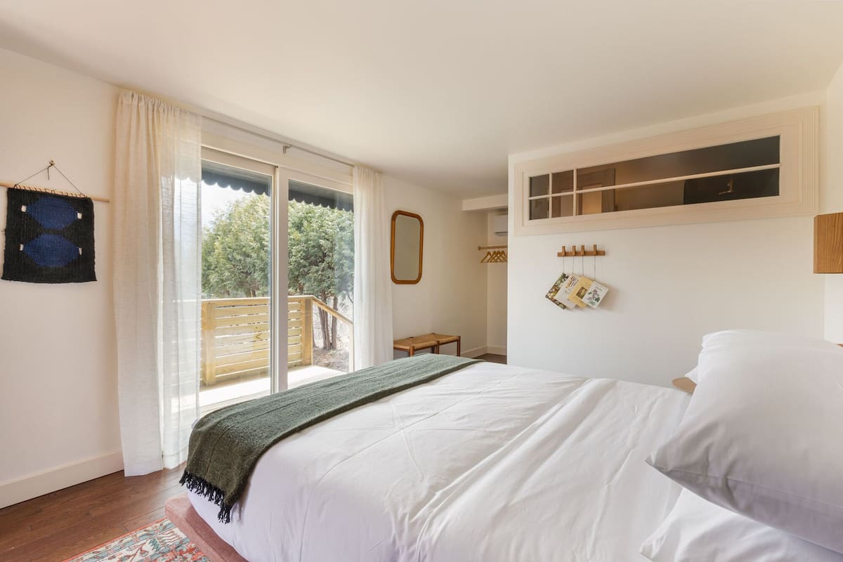 A bedroom with a large bed, a wooden bench, a small table, and a view of trees through a sliding glass door.
