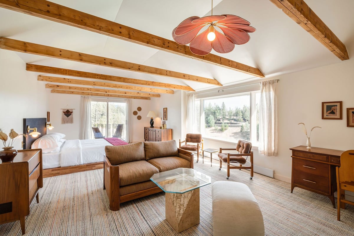A spacious room with a bed, a sofa, a glass coffee table, and a desk. There are large windows providing natural light and a view of the outdoors.