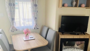 Une pièce avec une table en bois, deux chaises grises, un téléviseur posé sur un meuble en bois et une cheminée à la façade en pierre.