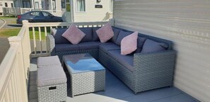 Ein überdachter Außenbereich mit einer grauen Rattan-Couch, zwei passenden Hocker und einem kleinen Tisch.