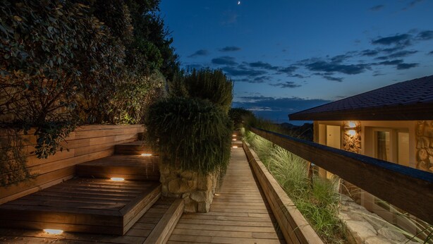 Un sentier en bois mène à un bâtiment en pierre surmonté d’un toit de tuiles, entouré de verdure et surplombé par un ciel nocturne nuageux.