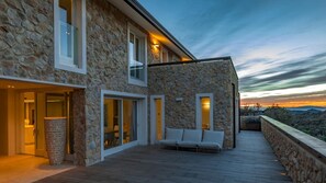 A modern stone building with large windows, a wooden deck, and a seating area. - Villa Le Mandrie 7 2 (Castiglione della Pescaia)