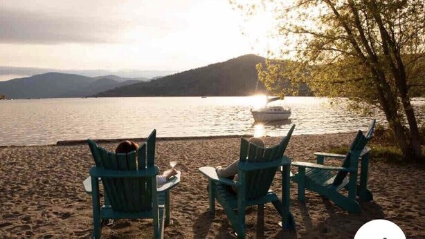 En strand med två gröna Adirondack-stolar som är placerade så att de vetter mot en sjö, och i fjärran syns en båt.