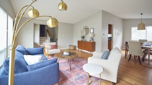 A spacious living room with a blue sofa, a white armchair, a round coffee table, and a patterned rug. A wooden cabinet and a dining area with a table and chairs are visible in the background. - Experience Woodinville Wine Country/Downtown Bothell (BOTHELL)