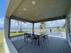 Un espace extérieur abrité, doté d’une table et de chaises, offrant une vue sur une plage et l’océan.