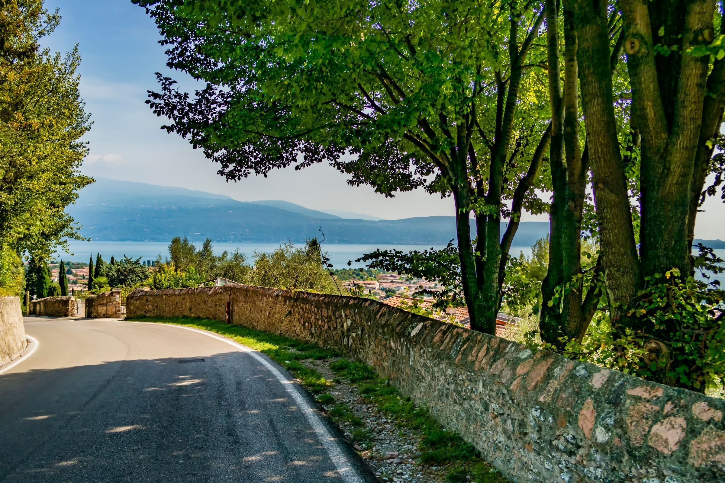 A curved road with stone walls on one side and trees on the other, leading to a view of a body of water and distant mountains.