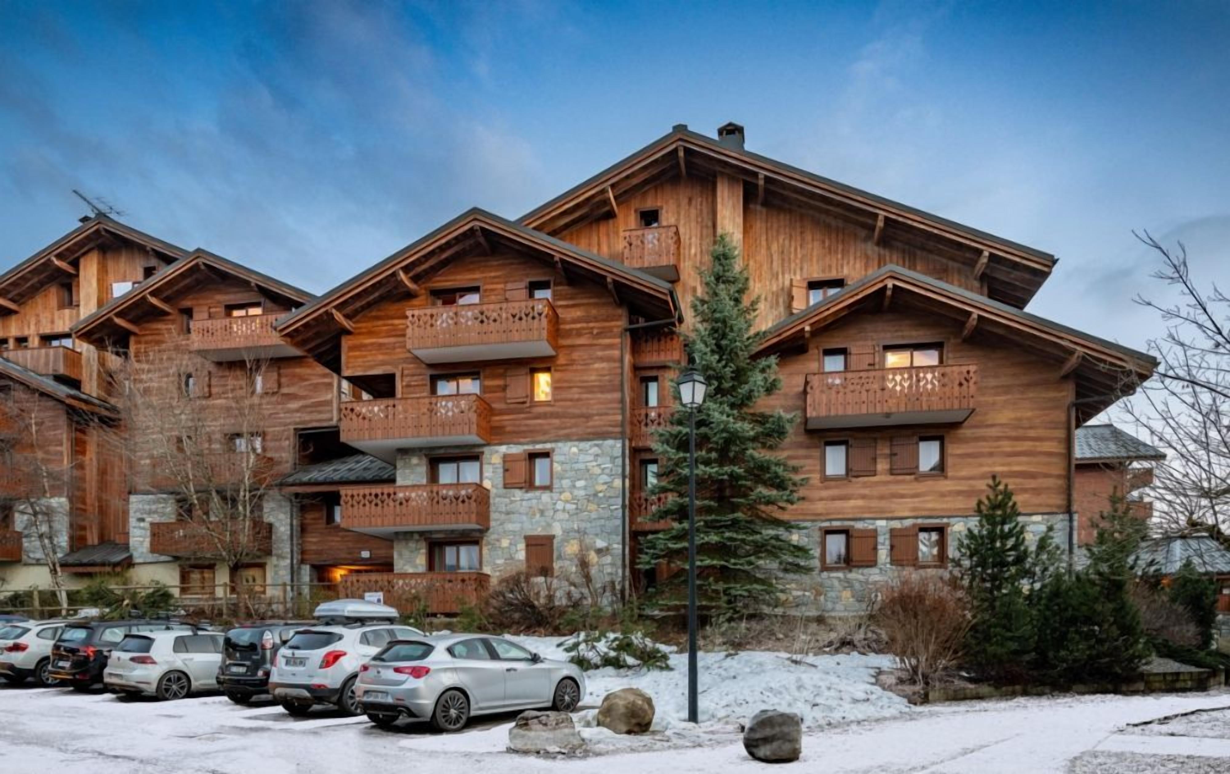 Un chalet en bois à plusieurs étages, doté de balcons, d’un espace de stationnement et surplombant un paysage enneigé.