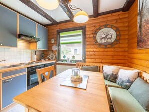 L’intérieur chaleureux d’un chalet avec une table à manger en bois, un banc intégré agrémenté de coussins, une cuisine aux armoires bleues et une grande fenêtre donnant sur un paysage verdoyant.