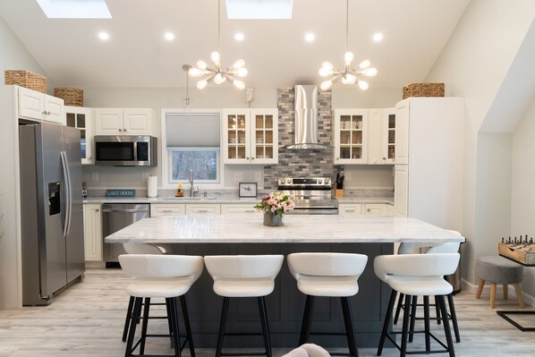 A modern kitchen with white cabinets, stainless steel appliances, a large island with bar stools, and decorative lighting.