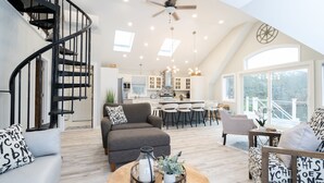 A spacious living area with a spiral staircase, a grey sofa, a round coffee table with decorative items, and a view of the kitchen and dining area through large windows. - New Lake House w/ Game Room & Stunning Views (Preston)
