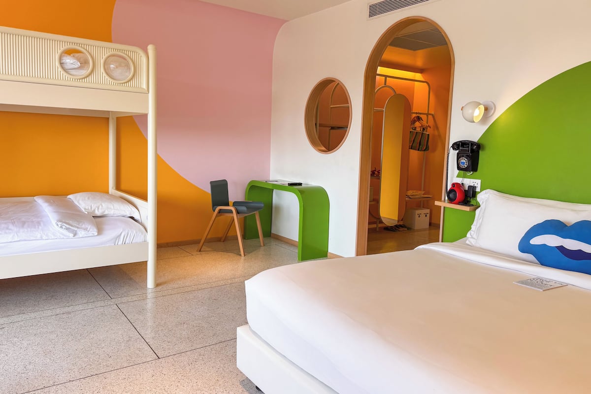 A hotel room with a bunk bed, a desk, a chair, a bed with a cloud design pillow, and a telephone.