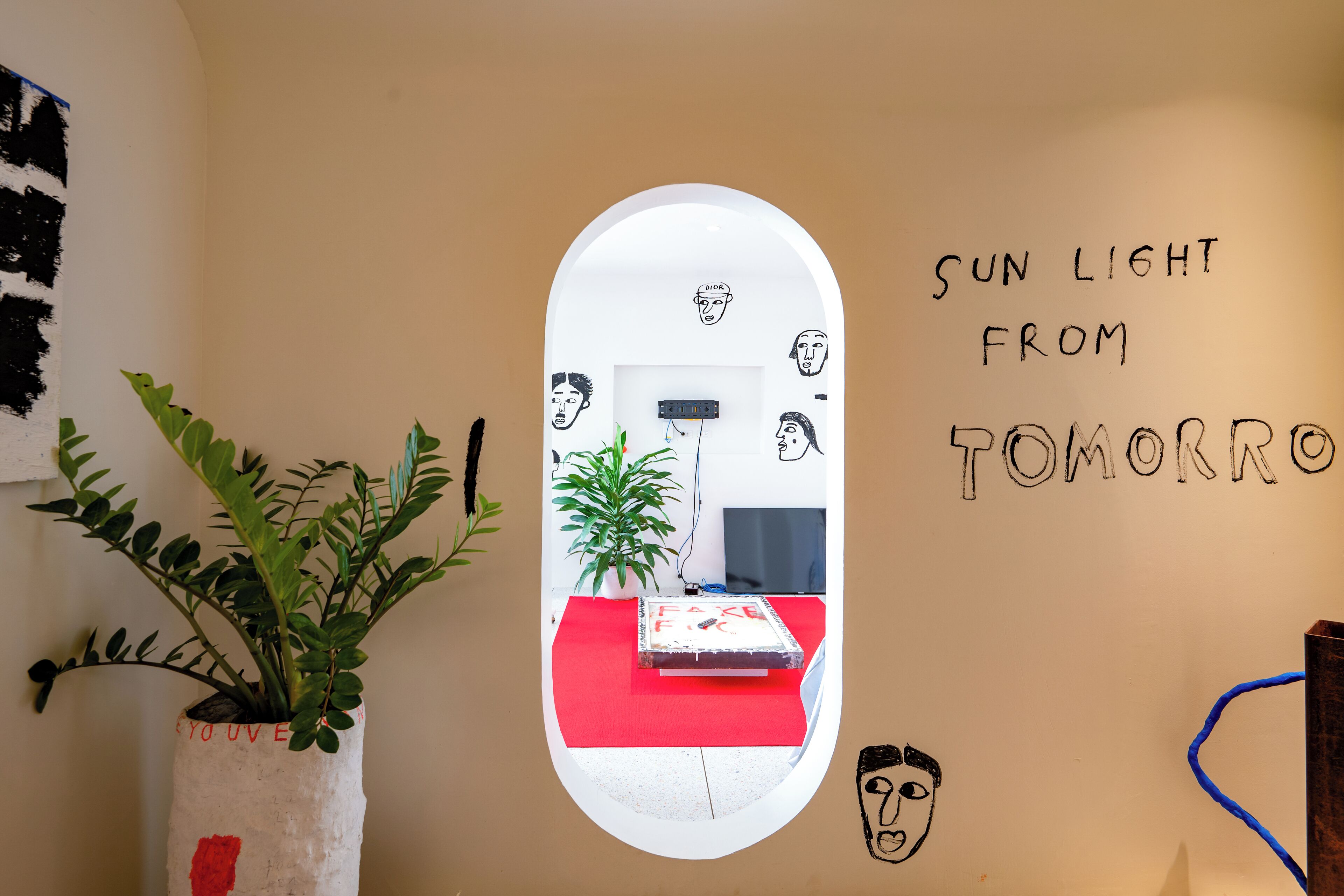 A room with a white wall, a mirror reflecting a red sofa and a plant, and a wall with text 'SUN LIGHT FROM TOMORROW'.
