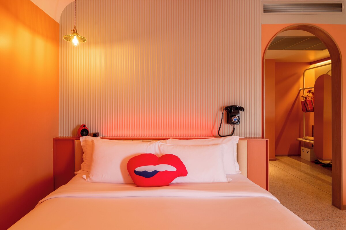 A hotel room with a bed, a red lip-shaped pillow, a telephone on the wall, and a corridor with a door.