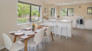 A modern kitchen with white cabinetry, a central island, and a dining area with white chairs and a wooden table. - Seas the Day - Matarangi Holiday Home (Matarangi)