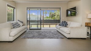 A living room with two sofas, a grey rug, a TV mounted on the wall, and a sliding glass door leading to a backyard. - Seas the Day - Matarangi Holiday Home (Matarangi)