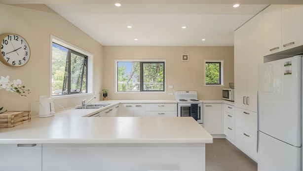 A modern kitchen with white cabinetry, a central island with stools, a stove, and a refrigerator. - Seas the Day - Matarangi Holiday Home (Matarangi)