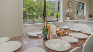 A dining table set with white plates, wine glasses, and a bottle of champagne. A window offers a view of trees and a fence. - Seas the Day - Matarangi Holiday Home (Matarangi)