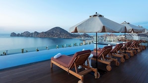 A poolside area with lounge chairs and umbrellas overlooking a body of water and a distant cityscape. - Corazon Ocean View Suite + Private Hot Tub • Sleeps 6 • Médano Beach • Dec 19–26 (Cabo San Lucas)