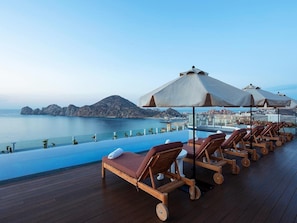 Ein Poolbereich mit Liegestühlen und Sonnenschirmen mit Blick auf ein Gewässer und eine ferne Skyline.