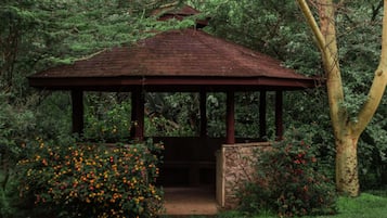 Ein Pavillon mit steinernen Fundament und Holzdach, umgeben von üppigem Grün und blühenden Büschen.