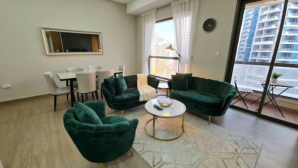 A modern living room with a dining area, green upholstered furniture, a round coffee table, and a large window offering a city view.