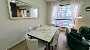 A dining area with a marble table, white chairs, a green sofa, and a large window with curtains.