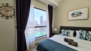 A hotel room with a large window, a bed with geometric patterned pillows, a clock on the wall, and a framed picture above the bed. - Marco Polo - Stunning Marina Views | High Floor | Close to Tram (Dubai)