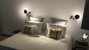 Cama bem arrumada com roupa de cama branca e almofadas listradas, mesa de cabeceira com abajur e arandelas na parede.