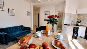 A dining area with a wooden table set for breakfast, including croissants, fruits, and a bottle of orange juice. A blue sofa and a chair are visible in the background. - Ze Perfect Place - Comfortable 2 Bedroom Apartment - Asc - Port - Garibaldi (Nice)