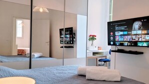 A modern hotel room with a large mirror, a flat-screen TV mounted on the wall, a bed with a blue blanket, a small desk with a chair, and a vase with red flowers. - Ze Perfect Place - Comfortable 2 Bedroom Apartment - Asc - Port - Garibaldi (Nice)