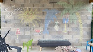 A room with a concrete block wall featuring colorful graffiti, a bench, a small table with a butterfly-patterned tablecloth, and a green chair. - Vacations in Pomié (Baladou)