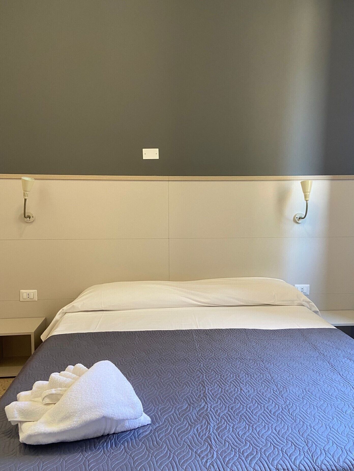 A neatly made bed with a blue quilt and white pillows, a white towel on the bed, and a wall-mounted light fixture.