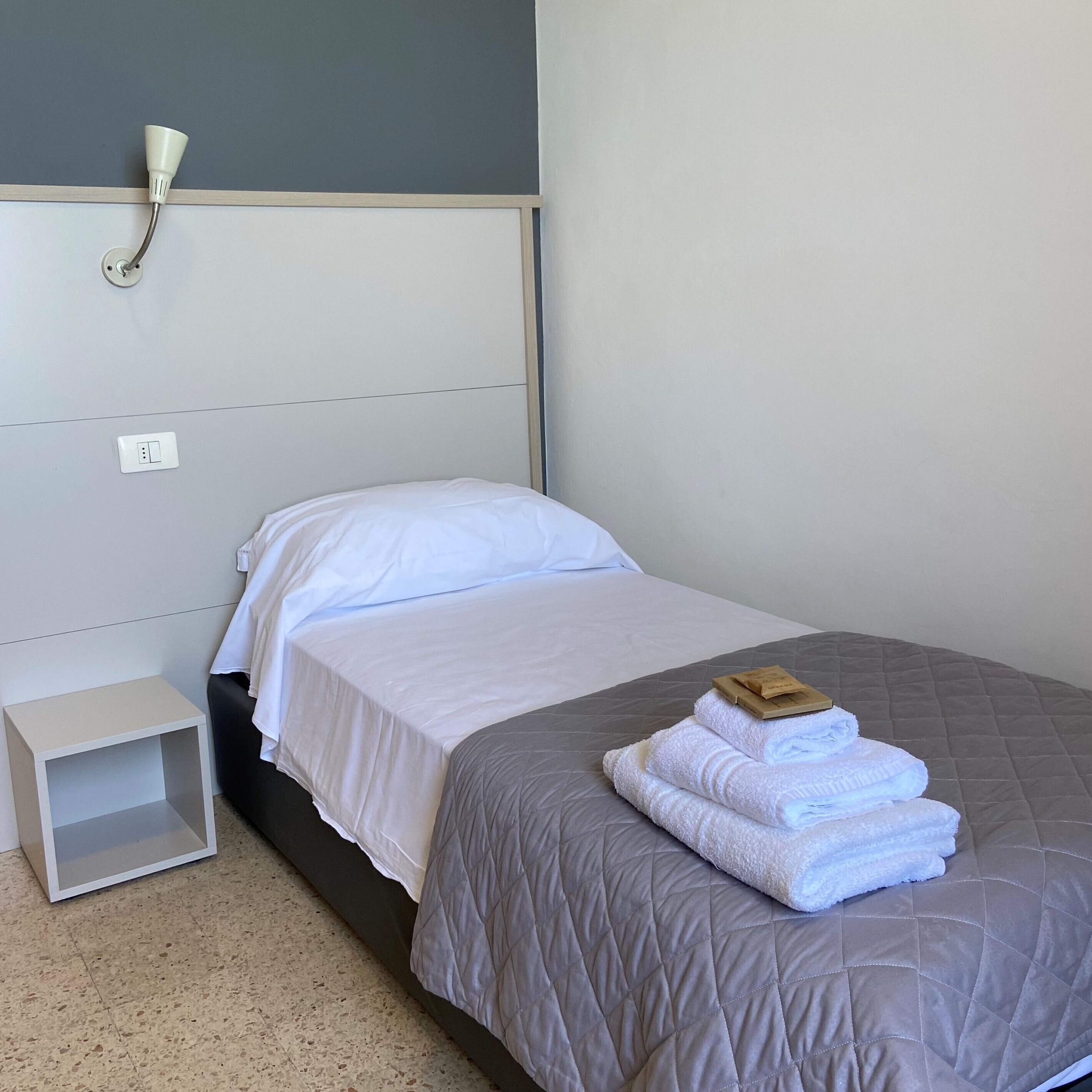 A neatly made bed with a grey quilt, a small bedside table, and a wall-mounted light fixture.