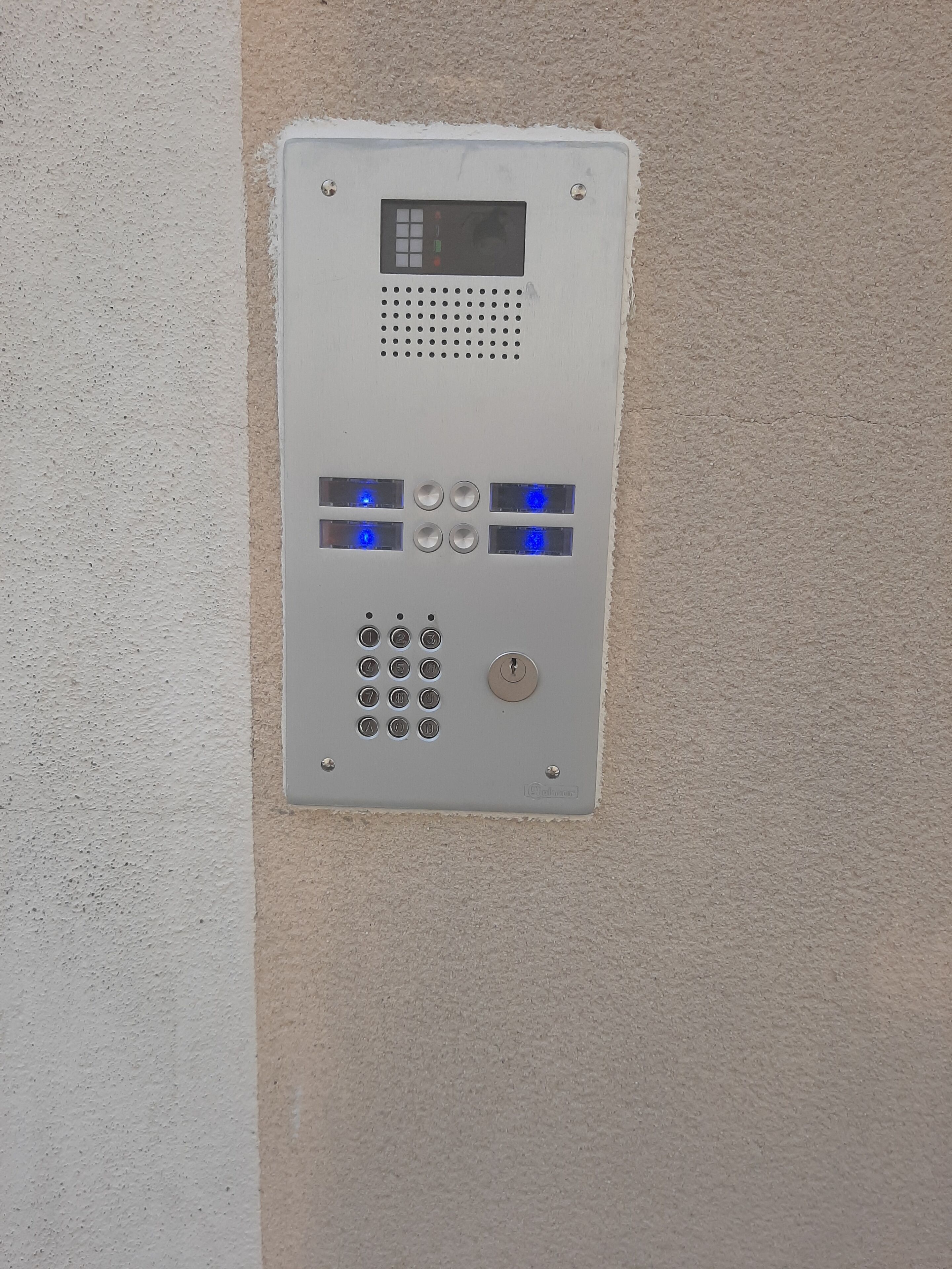 A wall-mounted intercom system with a keypad, a display screen, and illuminated buttons.