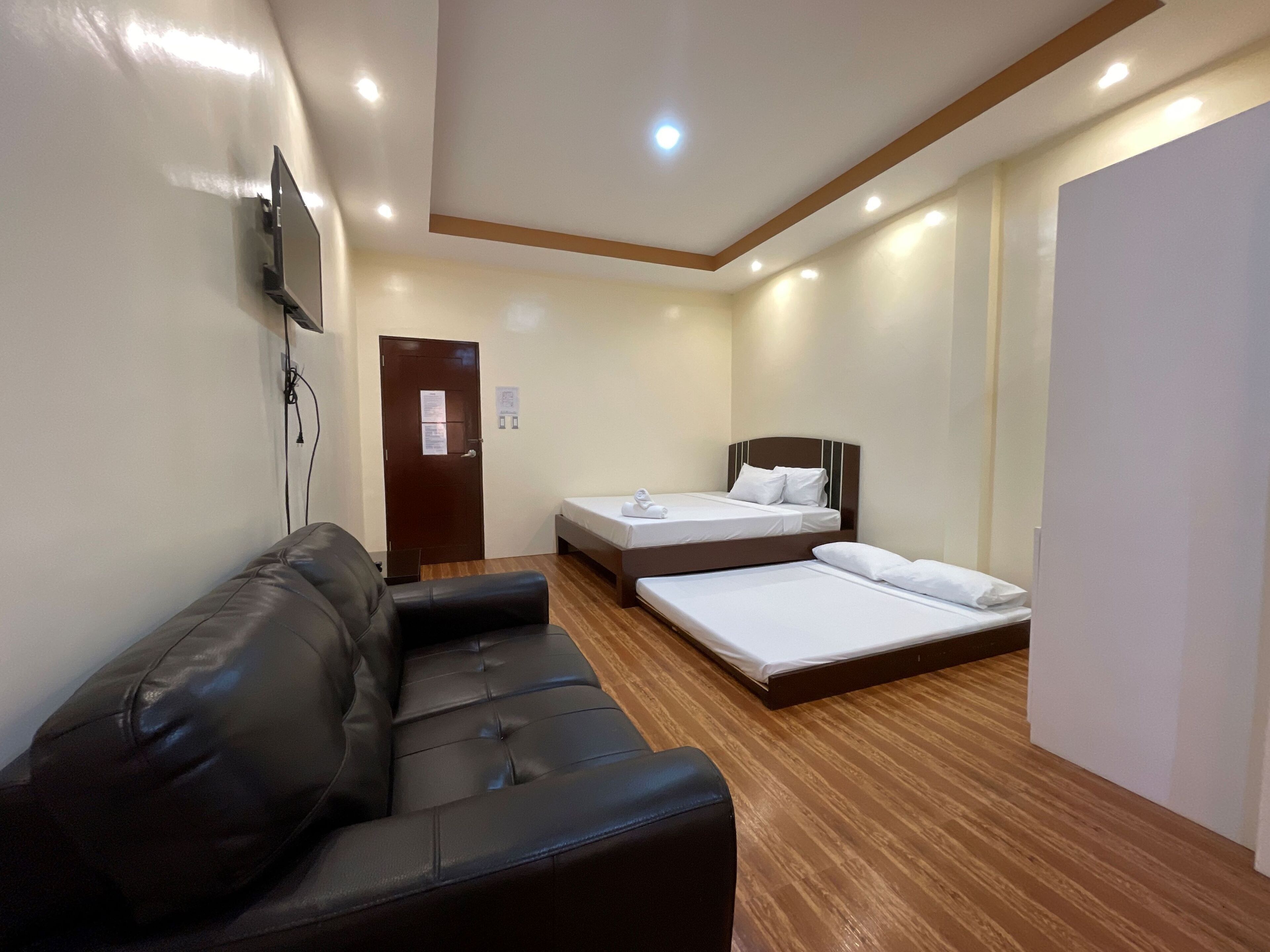 A hotel room with a black leather sofa, two single beds, wooden flooring, and recessed ceiling lighting.