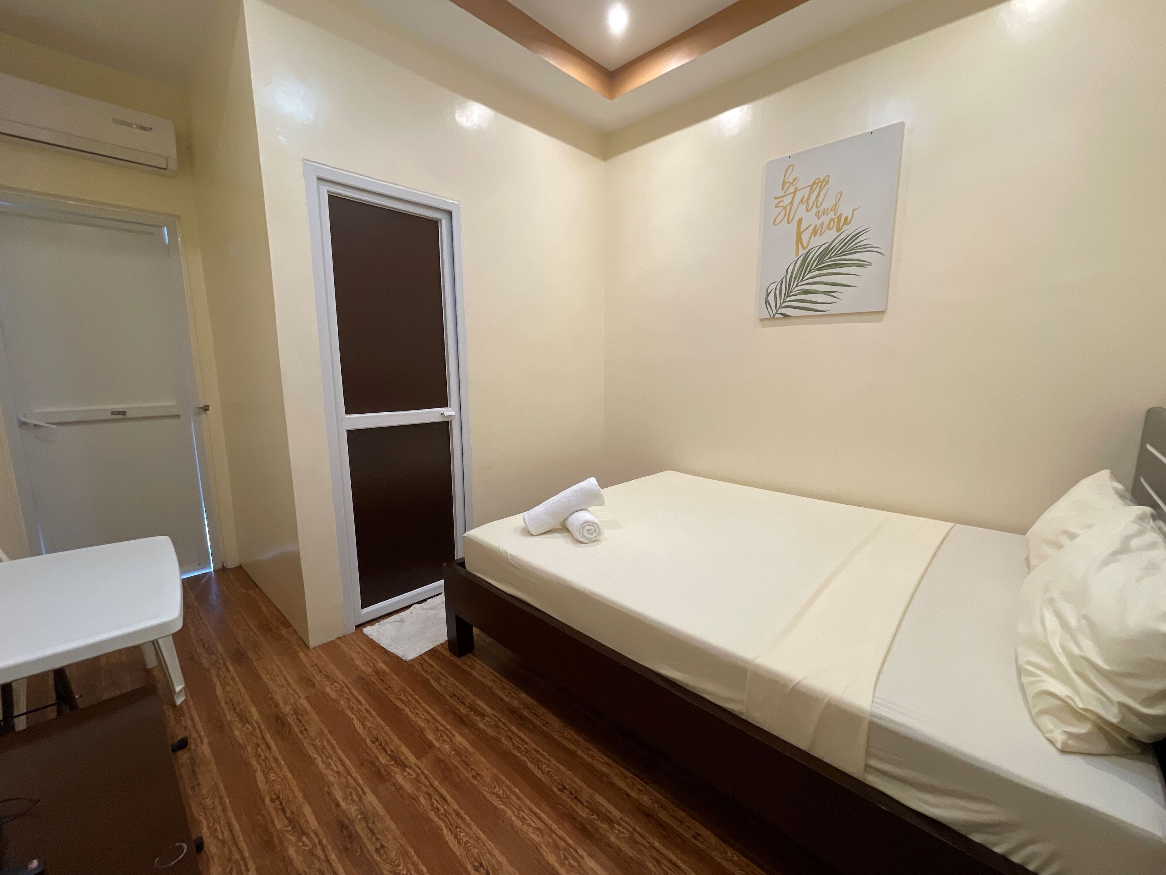 A small, neatly arranged bedroom with a single bed, a bedside table, a wardrobe, and a wall-mounted air conditioning unit.
