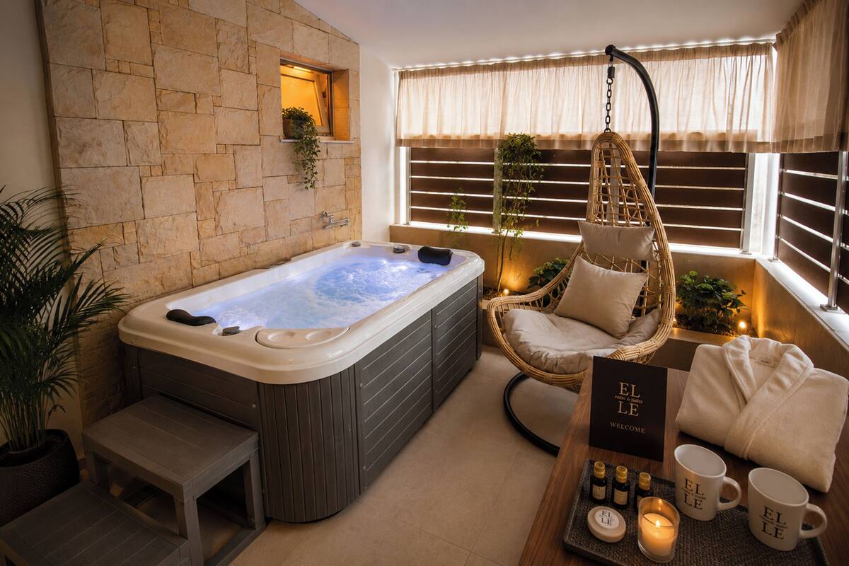 A spa room with a hot tub, a wicker chair, a bench, a tray with candles and a welcome sign, and a potted plant.