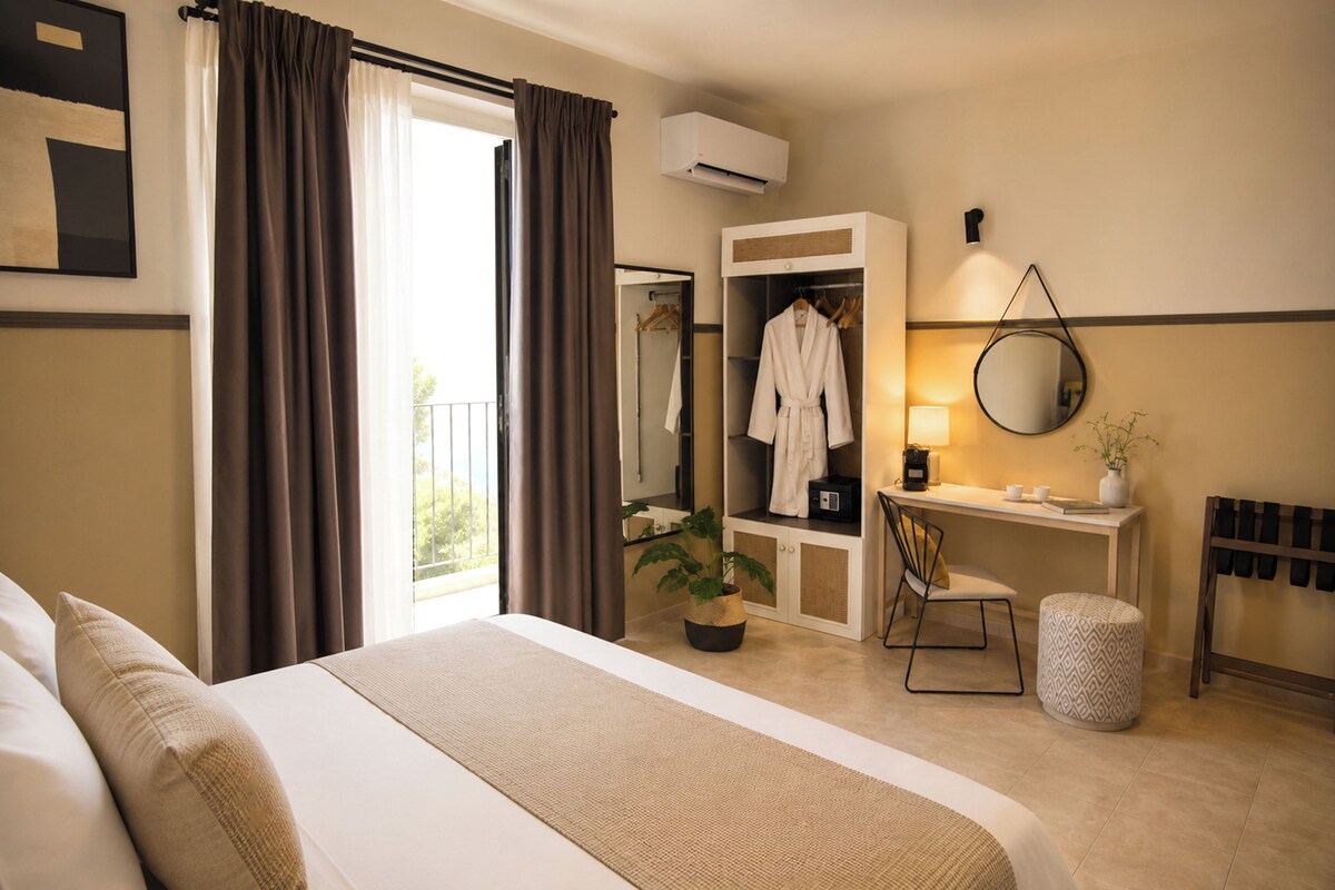 A hotel room with a bed, a desk with a mirror, a robe, and a potted plant.